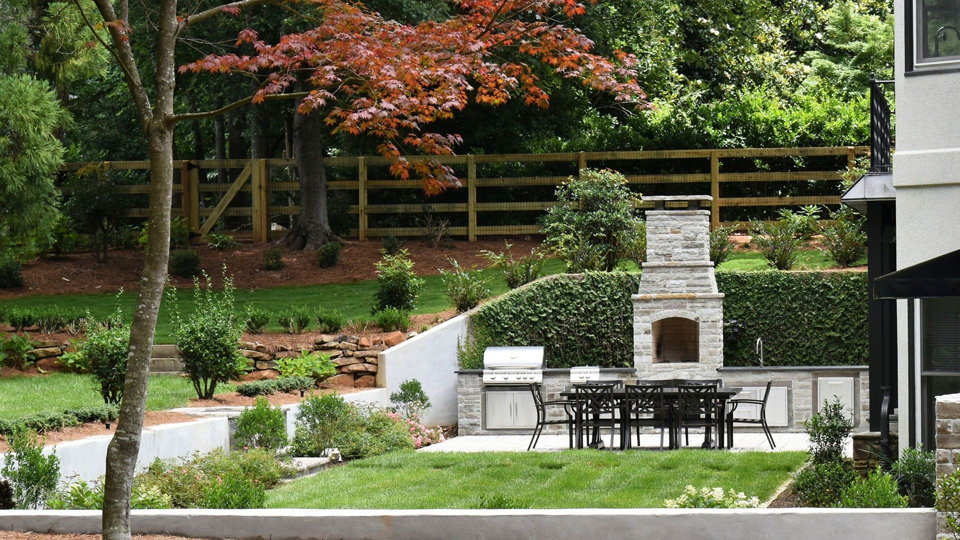 Outdoor patio with fireplace, grill, table, and greenery on a terraced lawn.