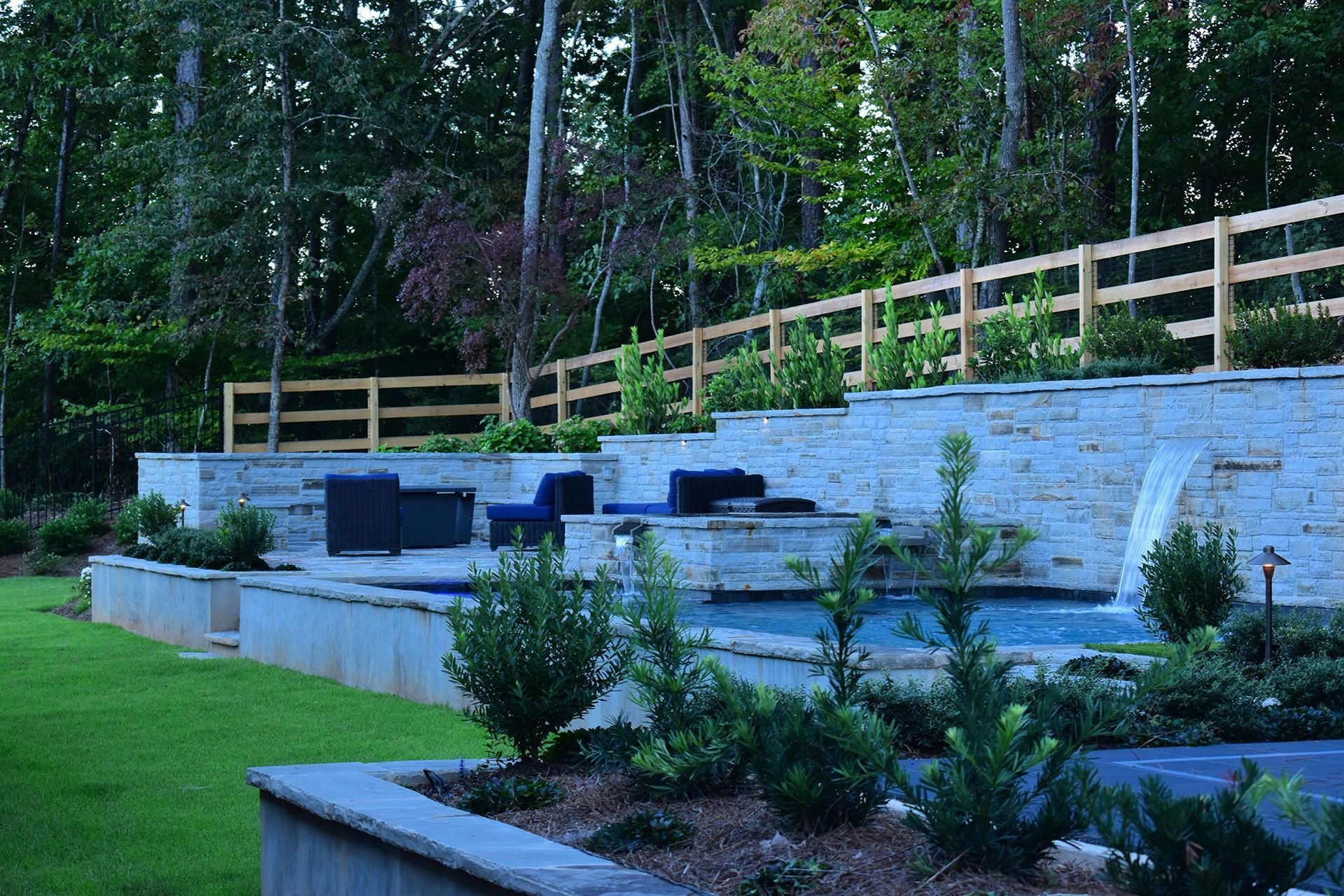 Multi-tiered backyard with pool, landscaping, and seating area, surrounded by trees and wooden fence.