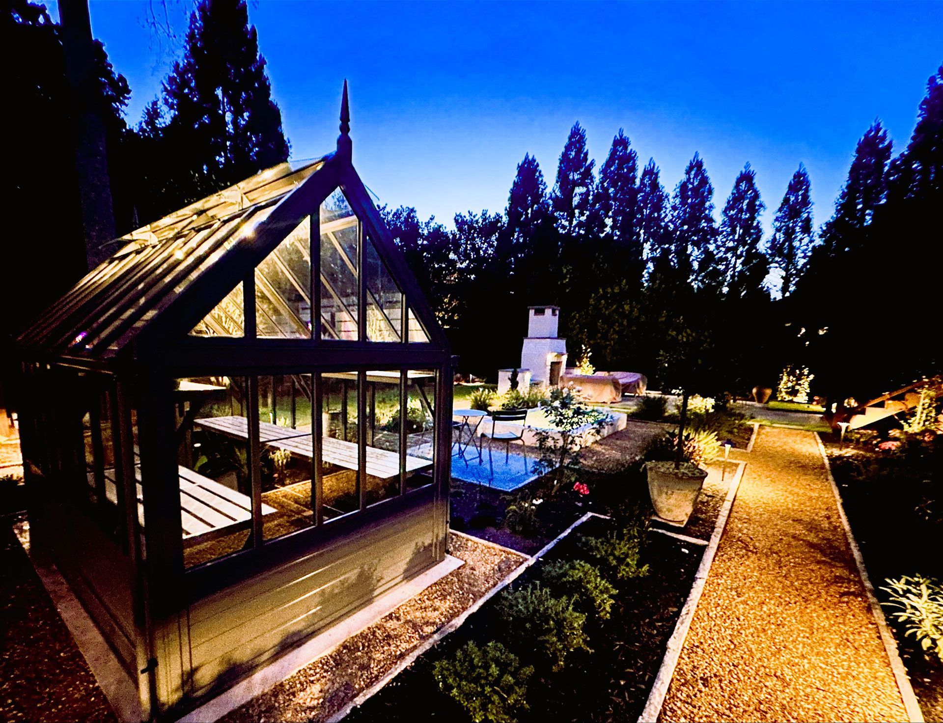 Greenhouse and garden illuminated at dusk. Pathways, pool, trees, and a distant white structure.