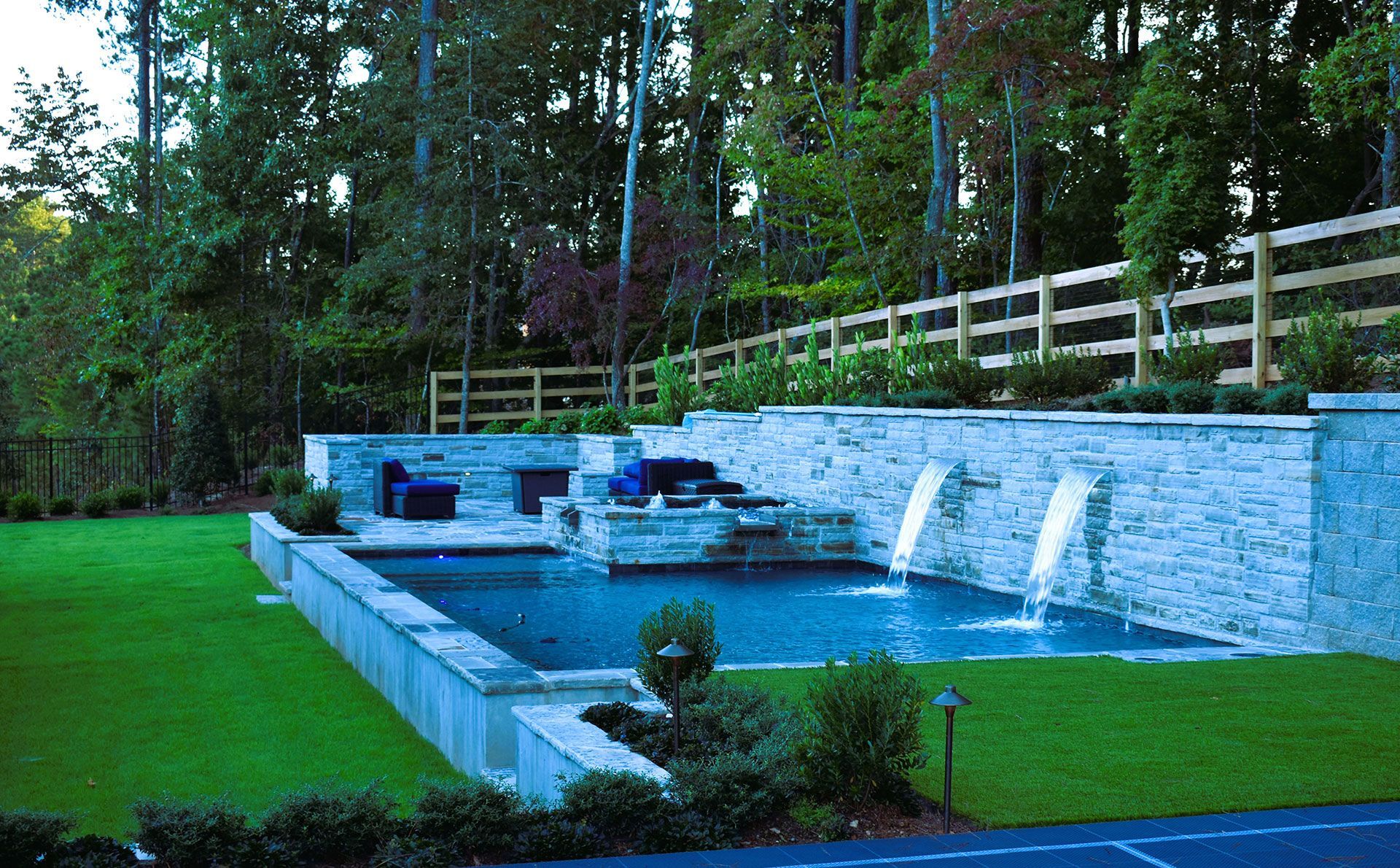 Modern pool with stone waterfall, surrounded by green lawn and trees. Wooden fence in the background.