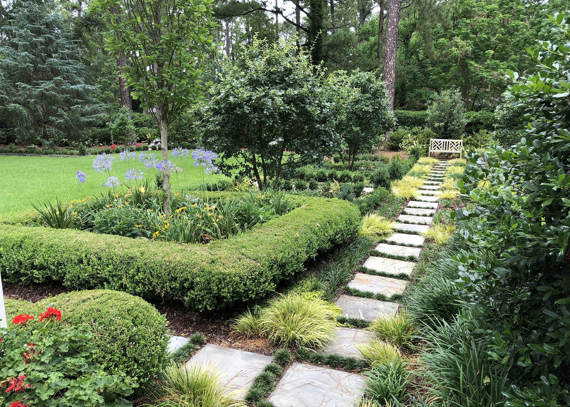 Stone path winds through a lush garden with hedges and flowers. Green foliage and trees surround the path.