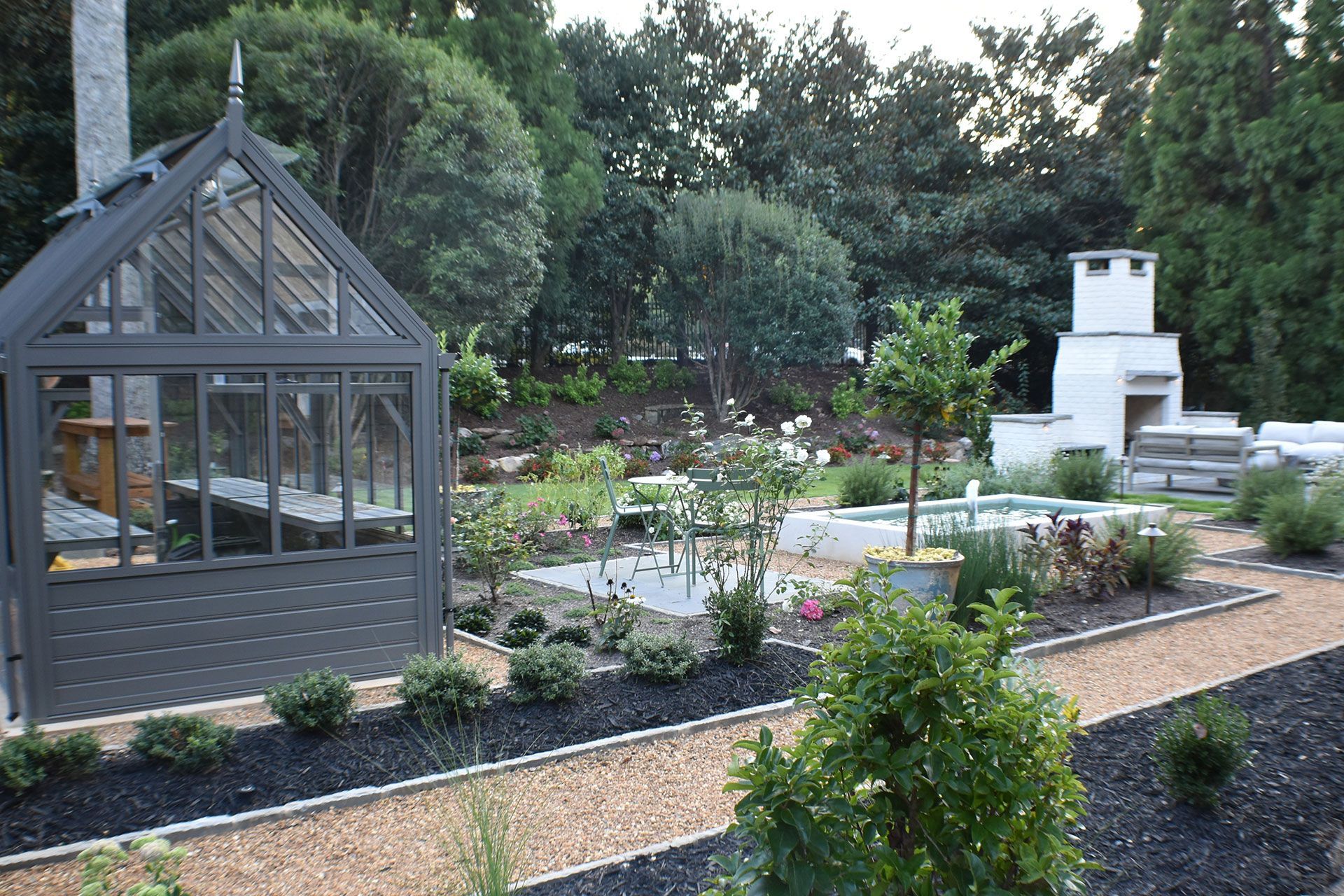 Greenhouse and garden with gravel paths, shrubs, and a white brick structure.