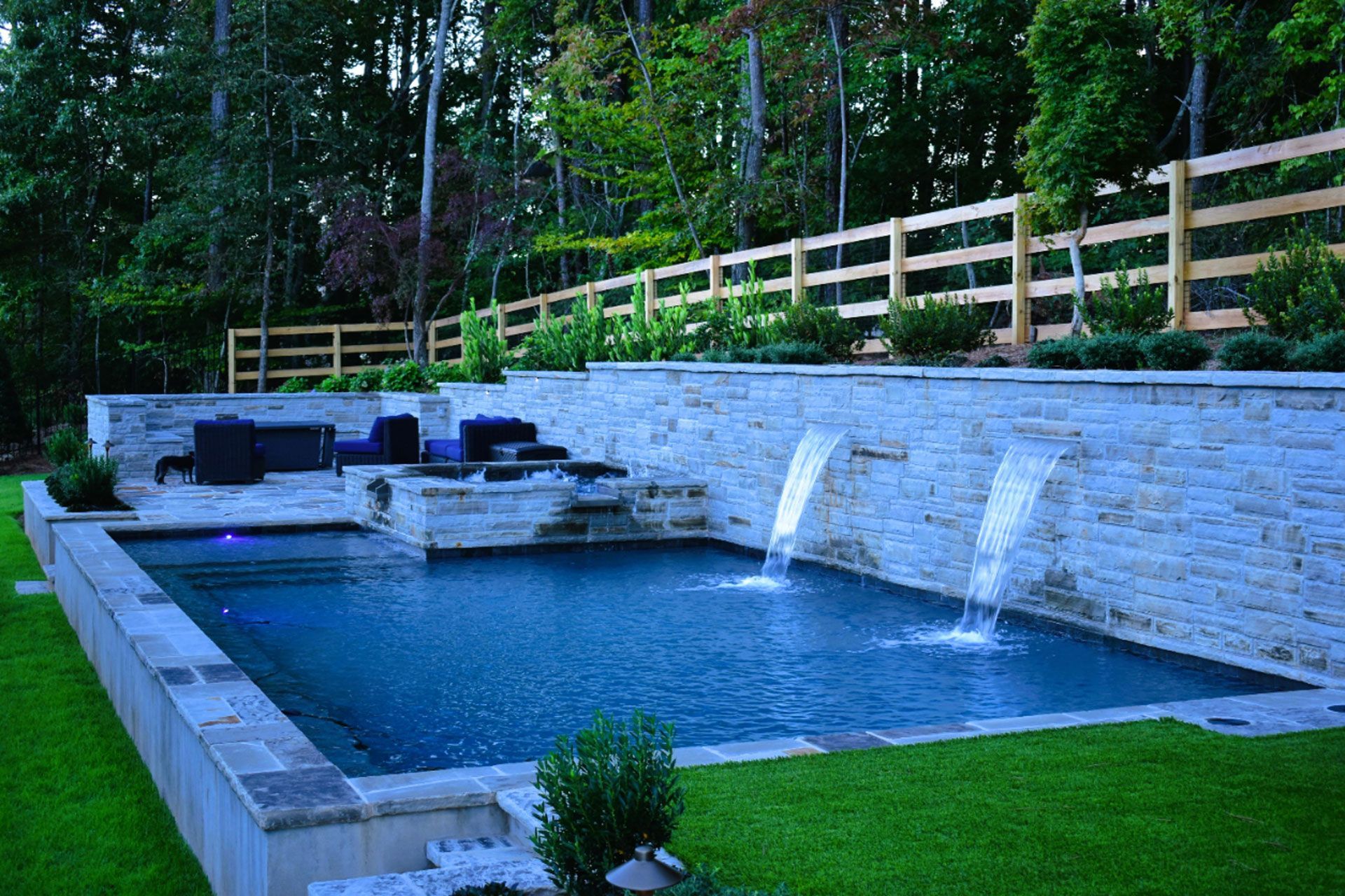 A luxurious backyard pool with waterfalls, a stone wall, and a wooden fence in a wooded area.