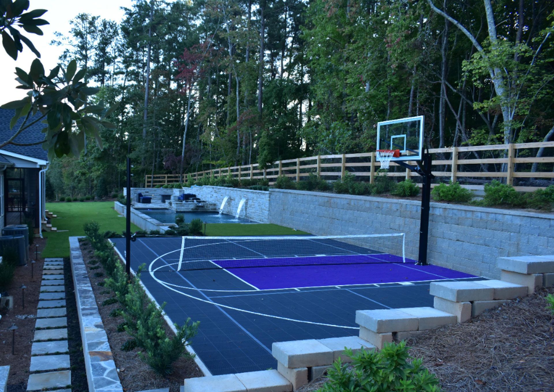 Outdoor basketball court with blue and gray surface. Green grass, pool, and trees in the background.