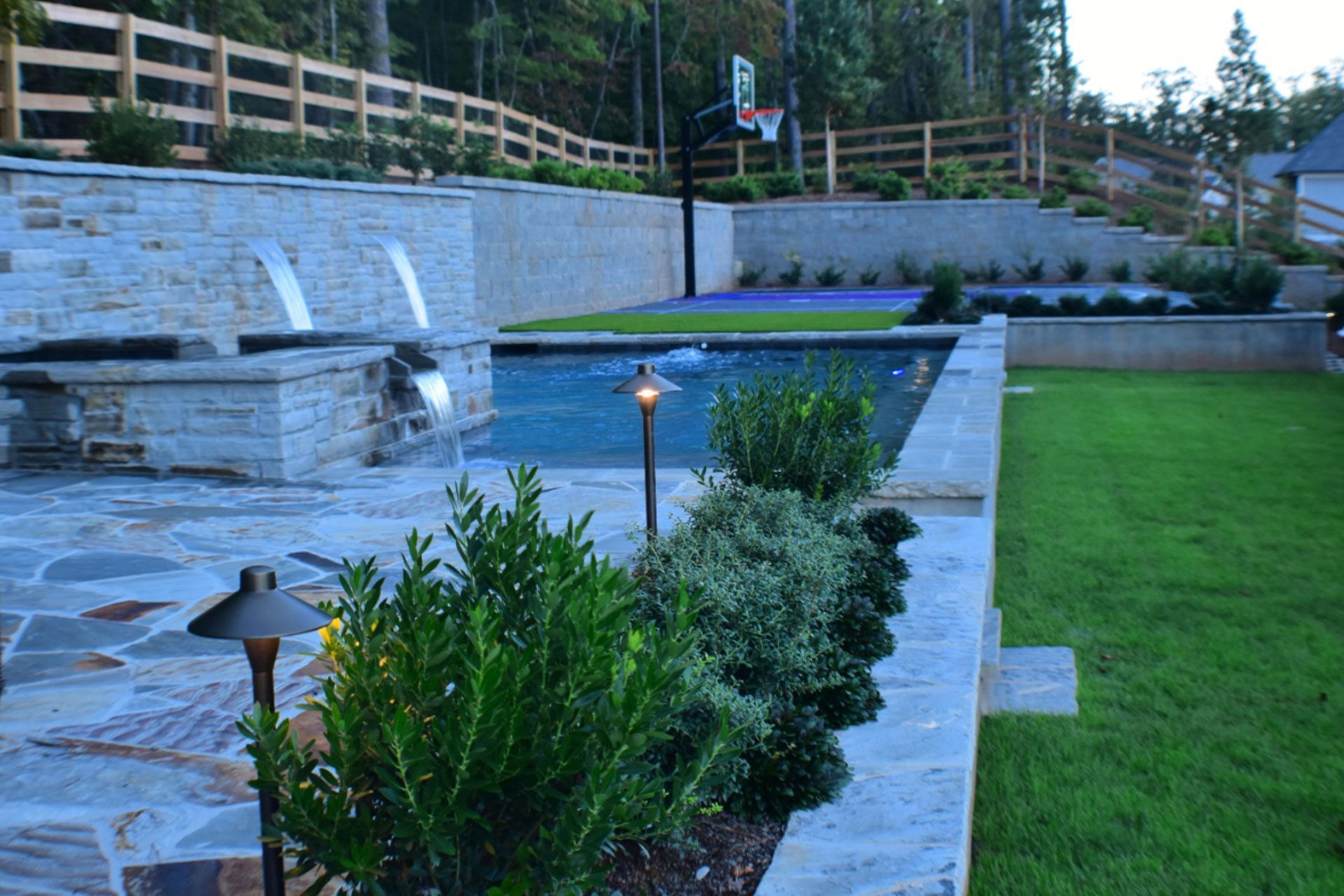 Outdoor pool and patio with stone features, a basketball hoop, and manicured landscaping.