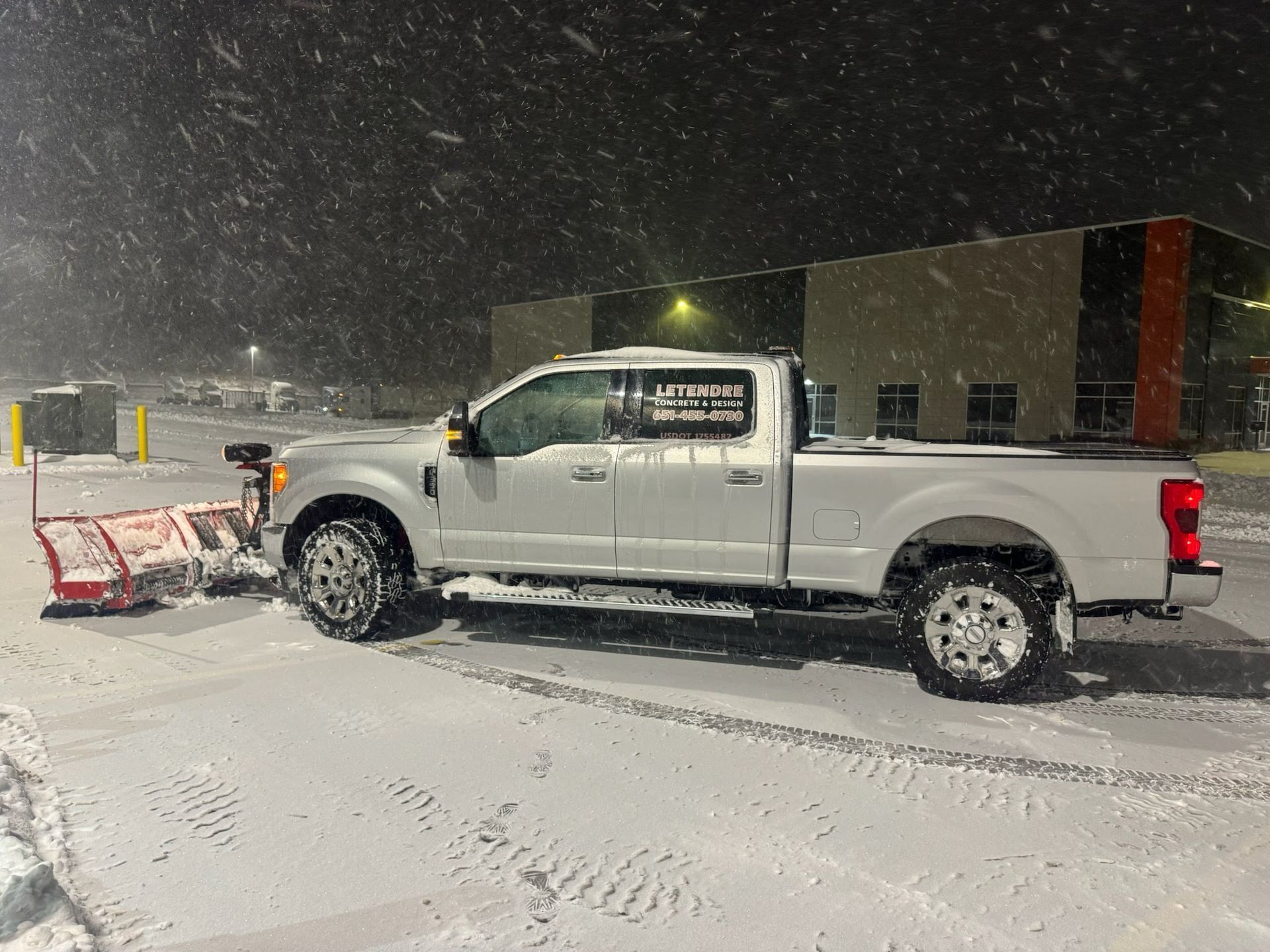 A silver pickup truck equipped with a snow plow clears a snowy parking lot at night.