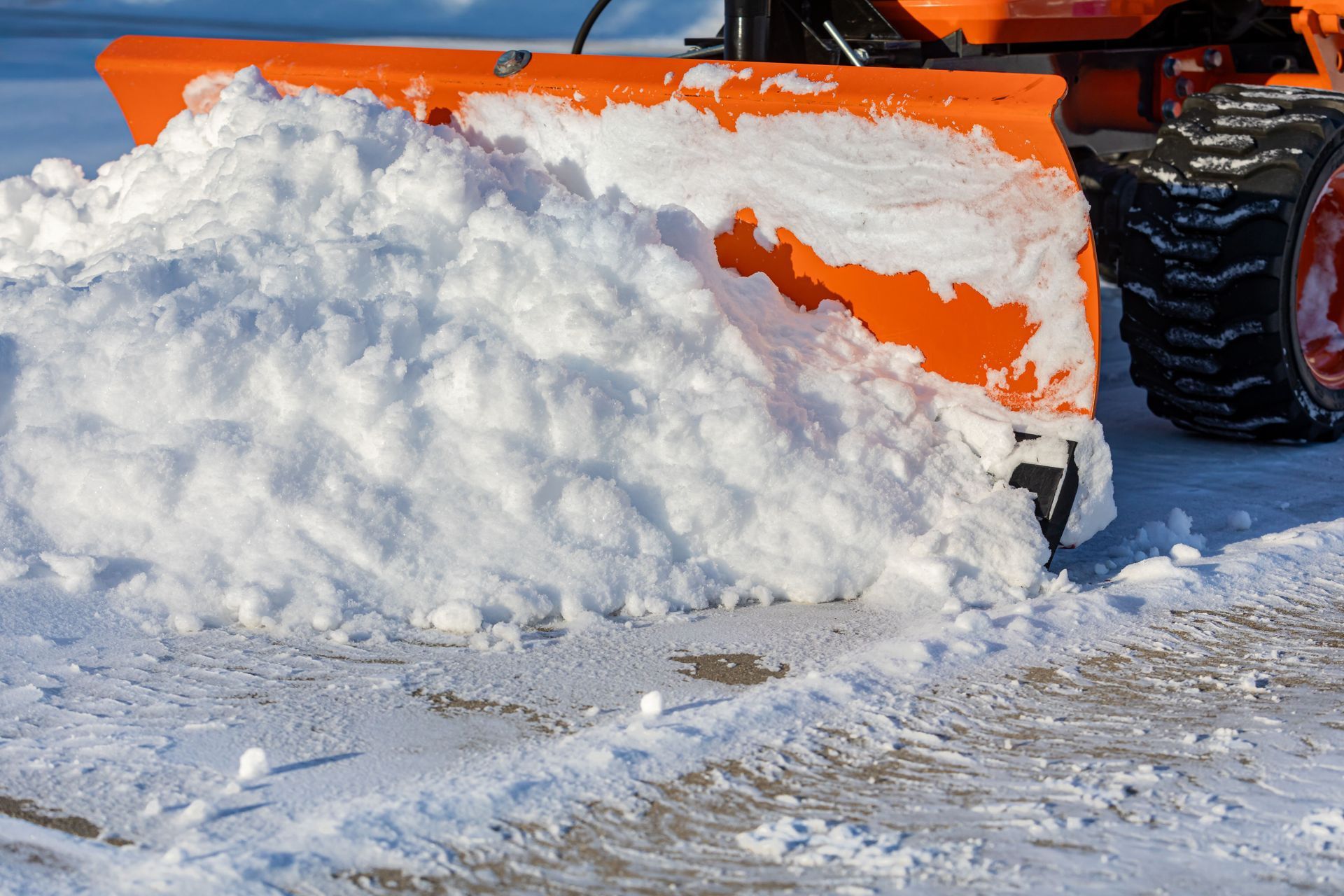 An orange snowplow blade pushing a large pile of white snow across a clear, icy ground surface.