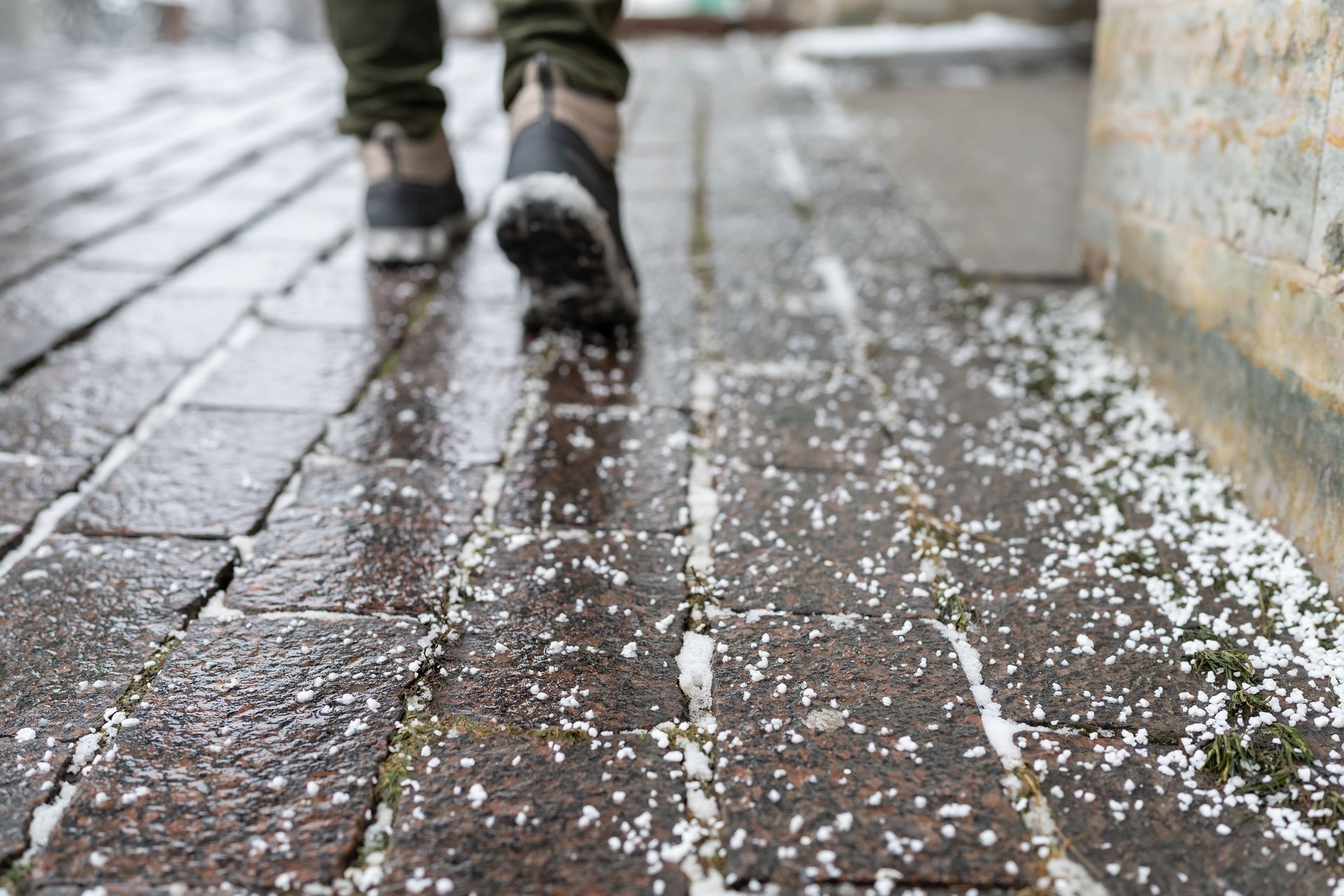 A person walks on wet, dark brick pavers covered with a light dusting of snow or ice.