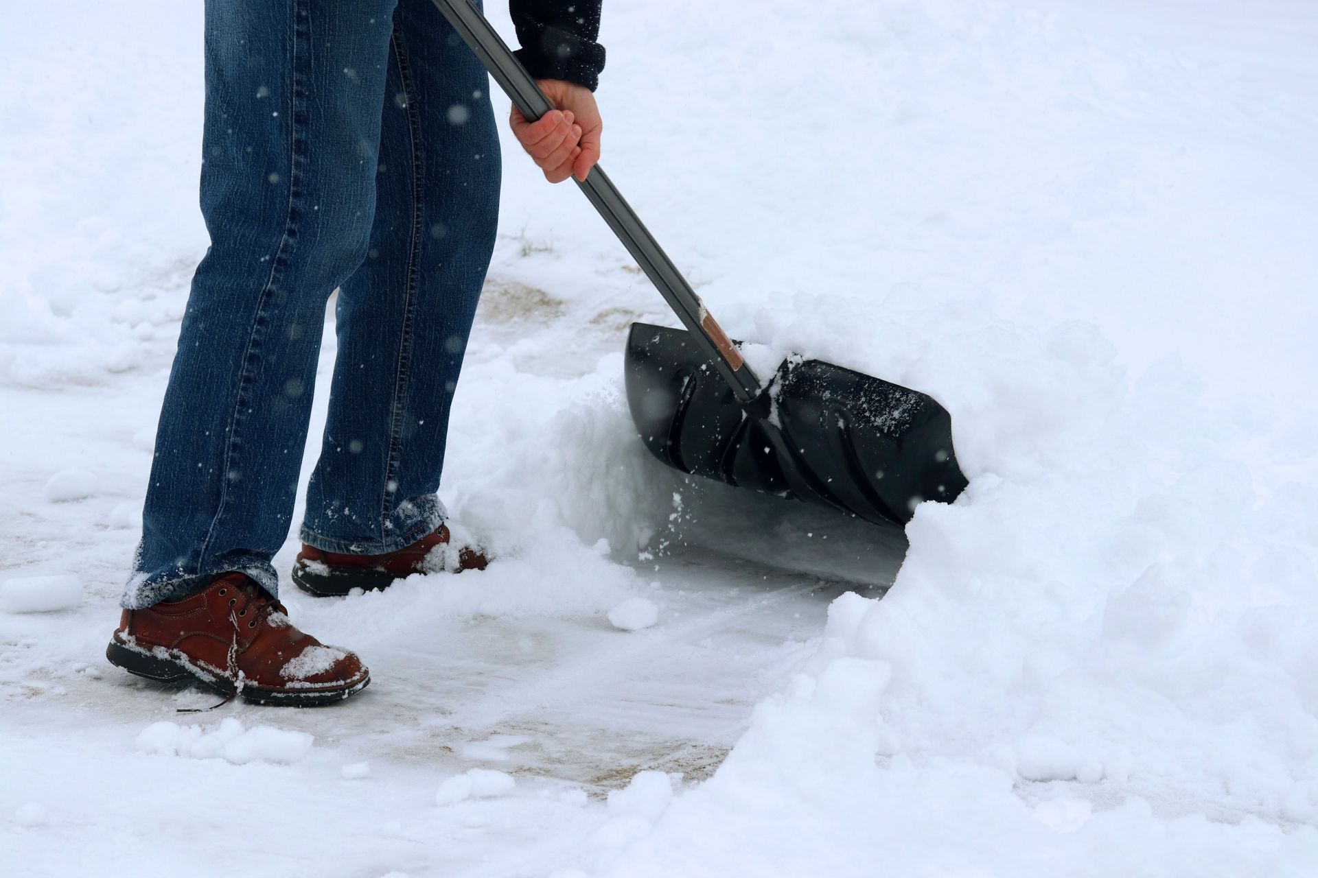 A person wearing blue jeans and brown boots uses a black shovel to clear snow from a ground surface.