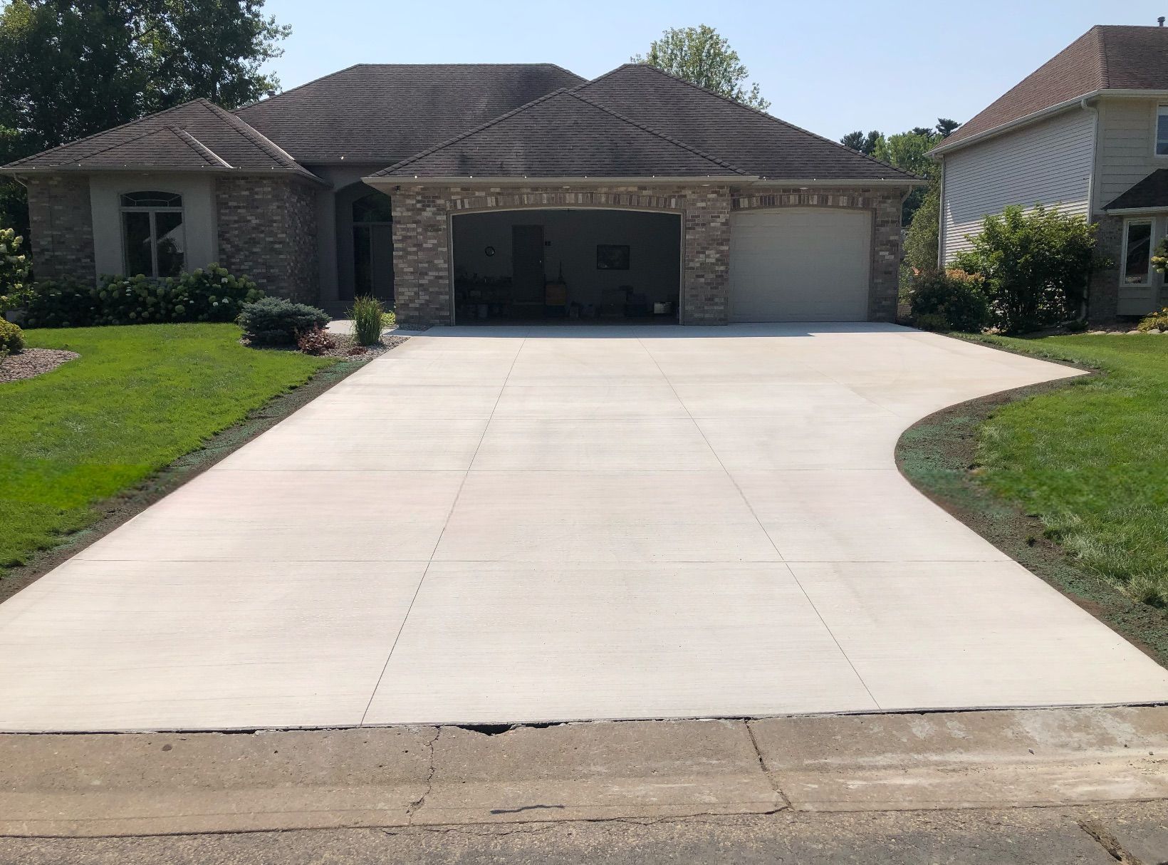 A new concrete driveway leads to the two-car garage of a single-story brick and stone house on a sunny day.