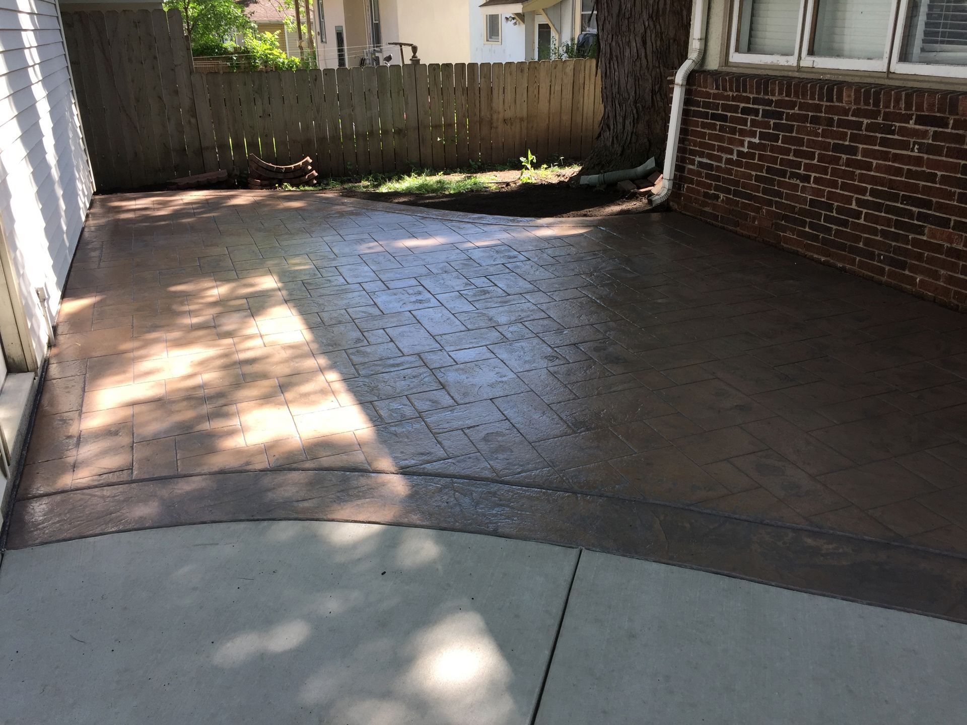 A stamped concrete patio featuring a grid pattern, transitioning from light brown to dark brown, next to a brick wall.
