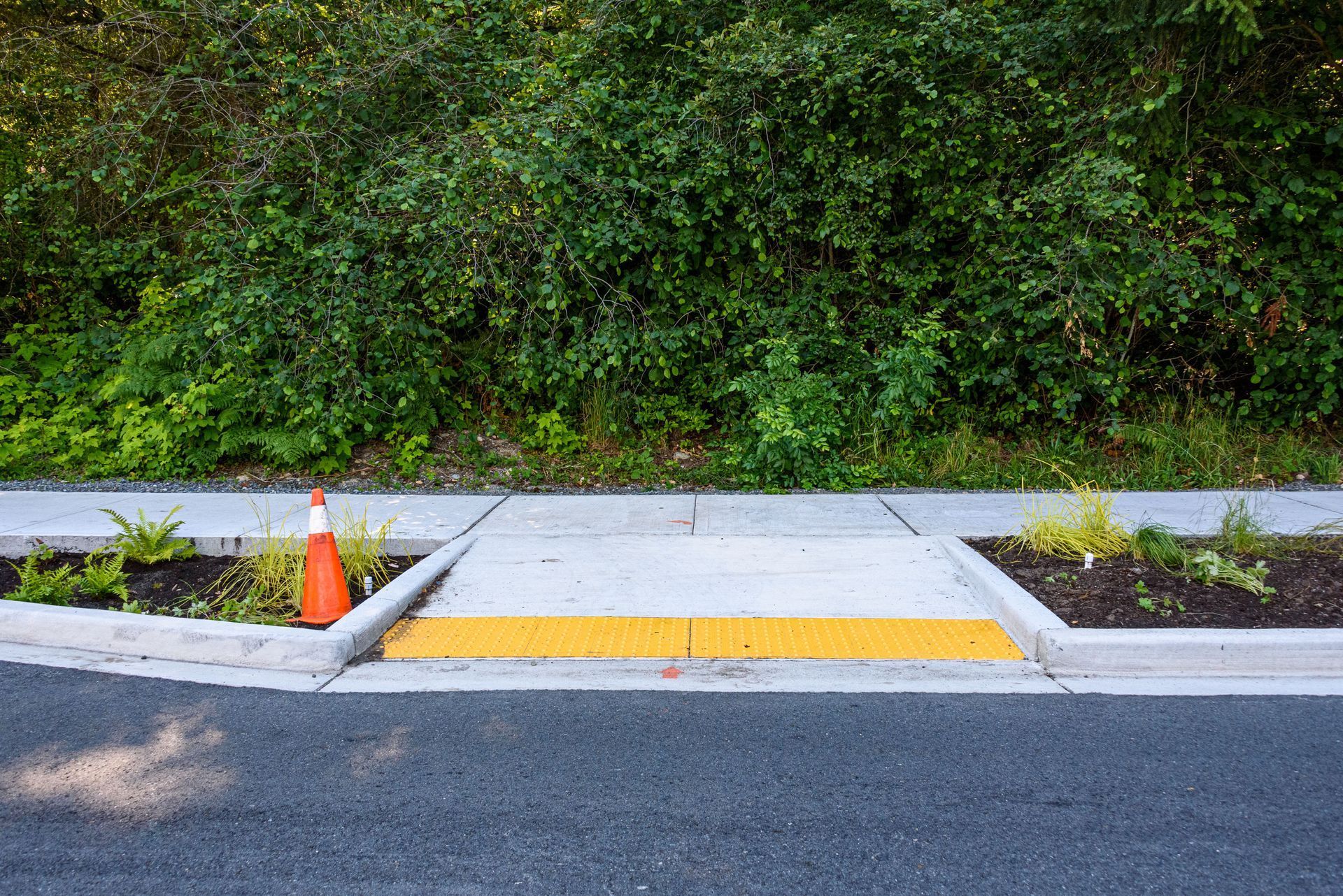 A concrete wheelchair ramp with yellow tactile paving on a curb, beside a small orange traffic cone.