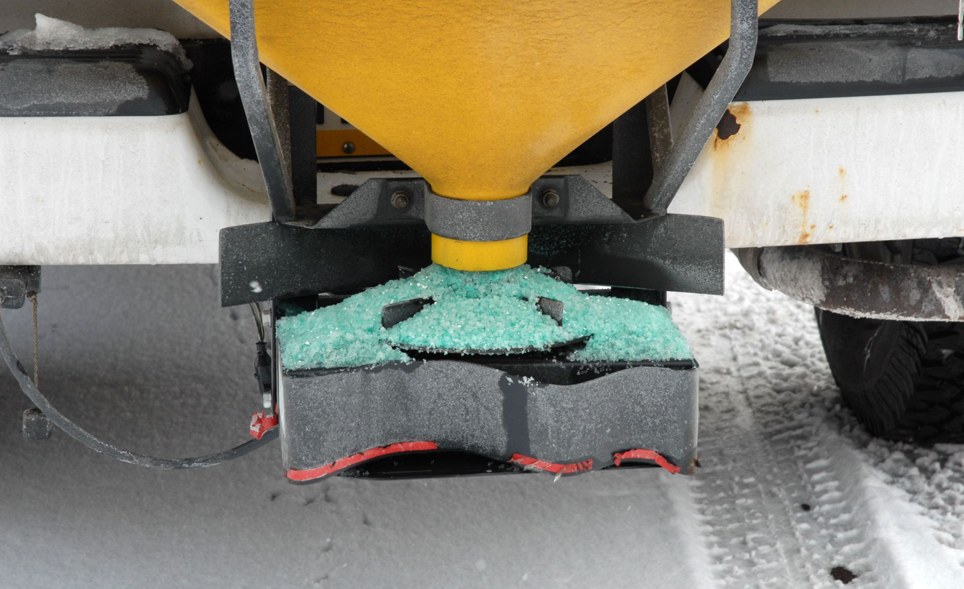 A snowplow salt spreader dispensing green salt onto a snowy road.