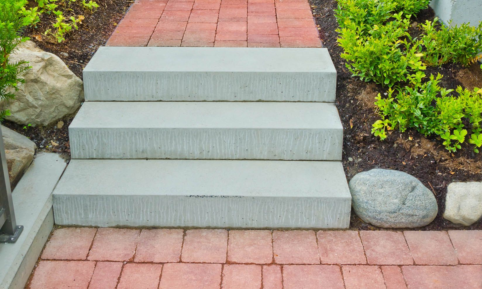 Red brick pathway with gray concrete steps leading upwards, flanked by greenery and brick wall.