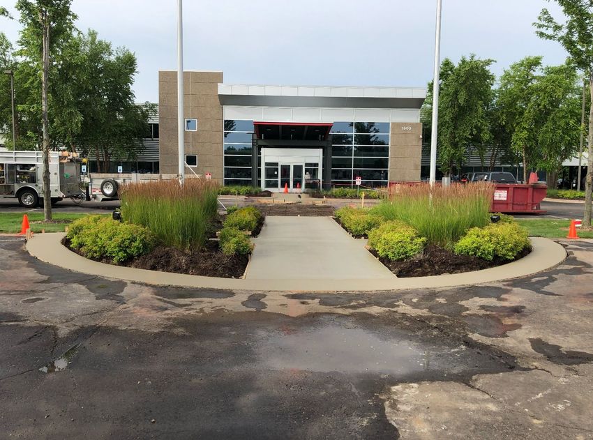 A modern commercial building entrance with a concrete path framed by circular garden beds in a parking lot.