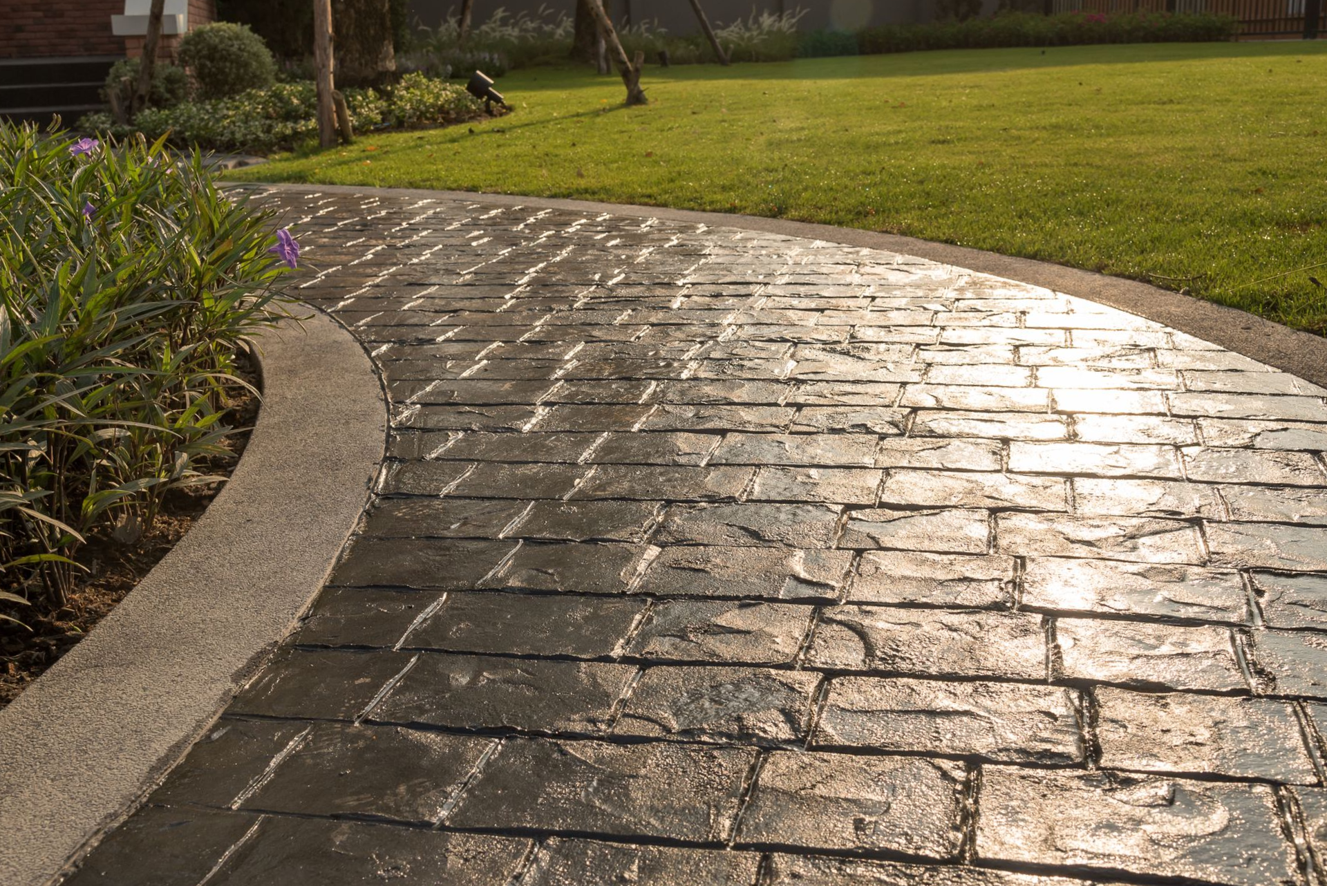 Brick-patterned walkway curving through a grassy lawn, bordered by plants and a concrete edge.