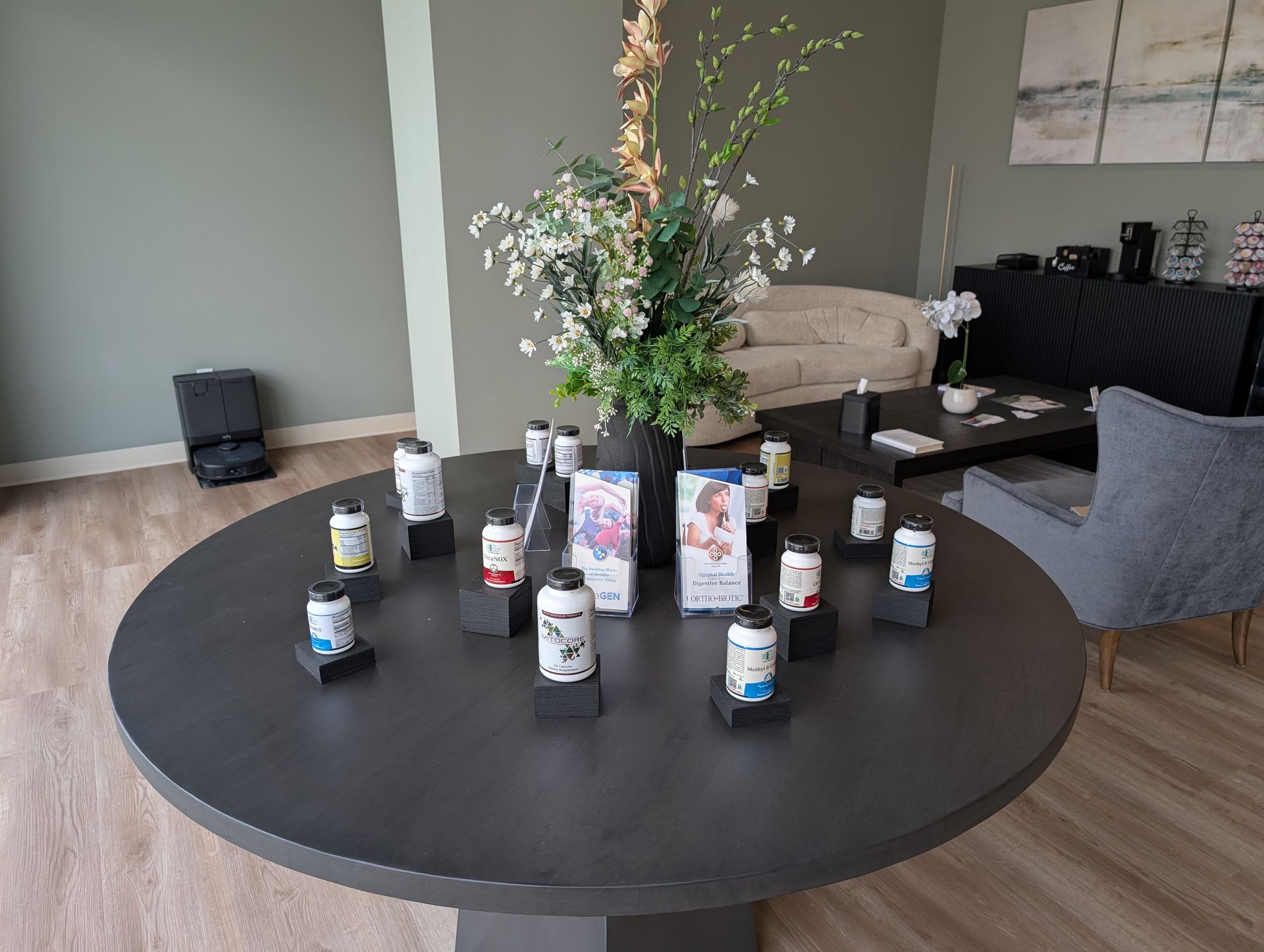 Round table displays supplement containers and promotional materials, decorated with flowers.