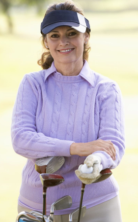 Woman wearing visor, holding golf clubs, smiling on a golf course. She's wearing a purple sweater and khaki pants.