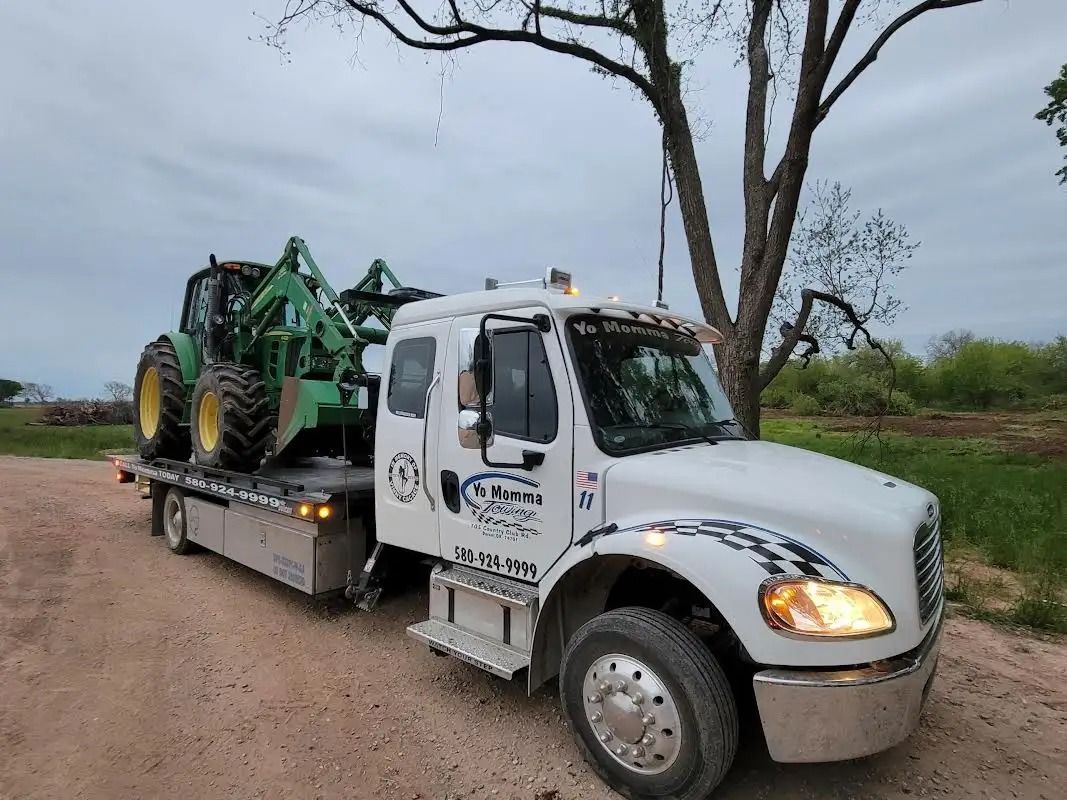 White tow truck hauling a green tractor on a dirt road, cloudy sky.