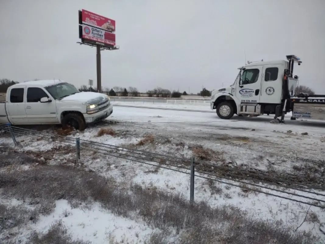 White truck off road with tow truck on snowy roadside near a billboard.