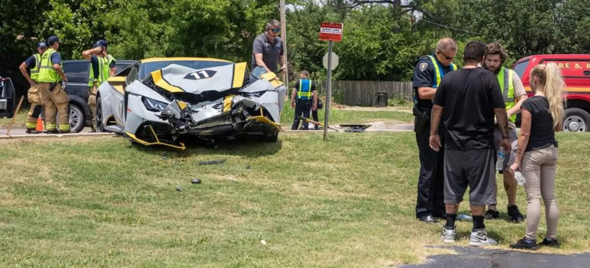 A damaged white sports car is on grass. People, including emergency responders, are assessing the scene.
