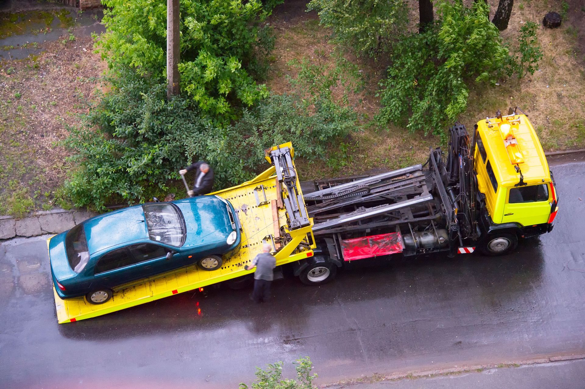 A tow truck loading a teal car on a rainy street, two people are present.