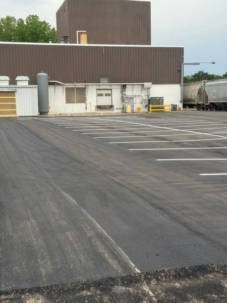 Black asphalt parking lot with painted white parking space lines in front of a brown and white industrial building.