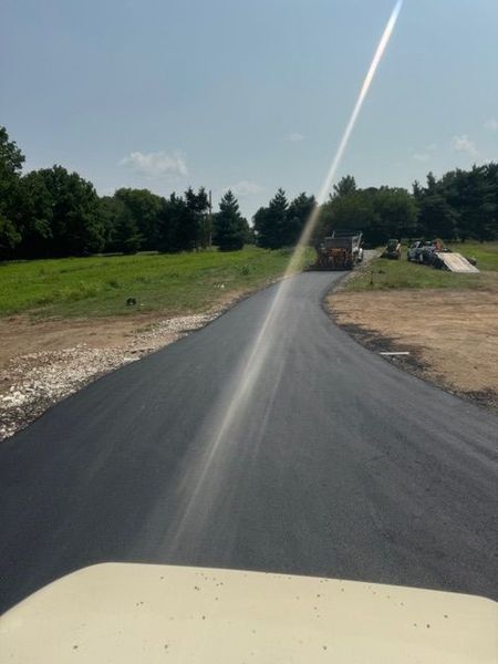 Newly paved asphalt road winding through a grassy field, under a bright, sunny sky.