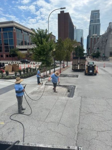 Three people spraying a city street with hoses. Buildings and a small vehicle are visible.