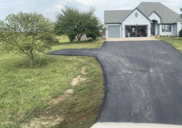 Black asphalt driveway leads to a light blue house with a two-car garage. Green grass surrounds the driveway.