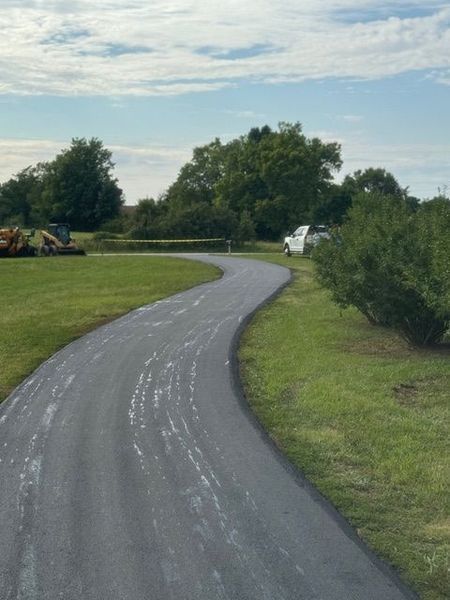 Newly paved asphalt driveway curves through a grassy lawn, trees in the background, a truck parked ahead.