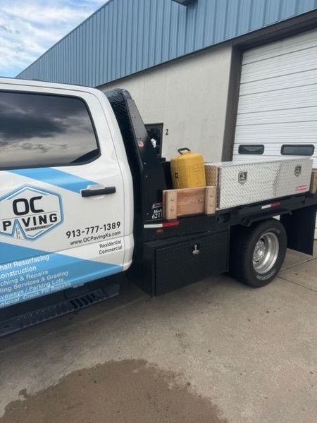 White work truck with OC Paving logo parked by a building, bed holds tools and equipment.