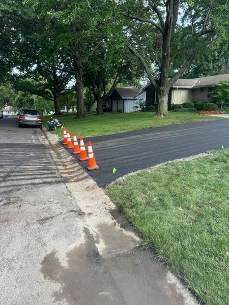 New asphalt driveway with orange cones on the edge, next to a cracked sidewalk and green grass.