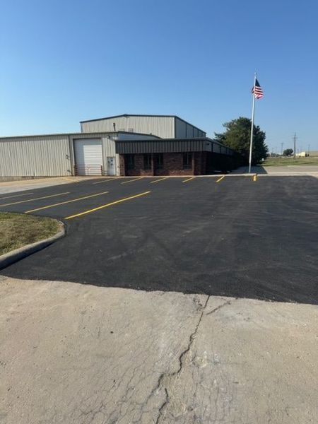 Black-paved parking lot in front of a low, tan building with an American flag on a pole under a clear blue sky.