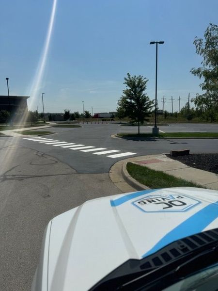 Crosswalk in a parking lot on a sunny day. White zebra stripes lead to a grassy area and building.