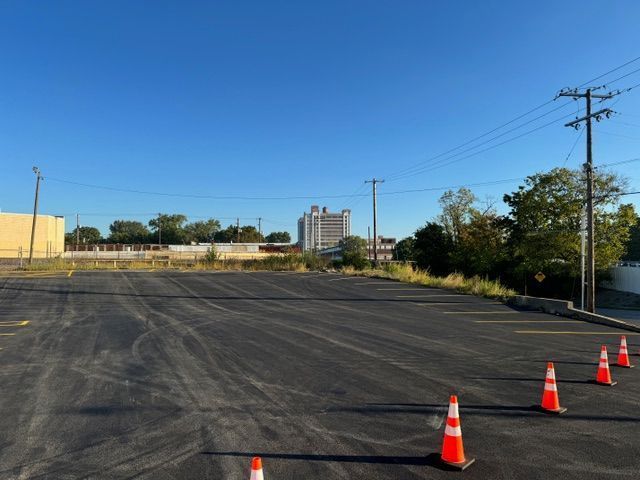 Empty asphalt parking lot with orange cones, buildings in the background. Clear blue sky.