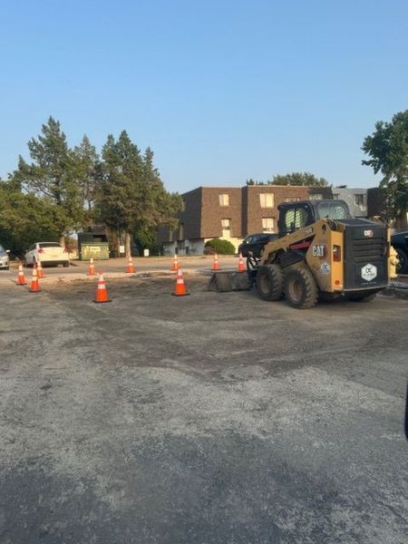 A skid steer operating in a parking lot, surrounded by orange cones. Asphalt is being worked.