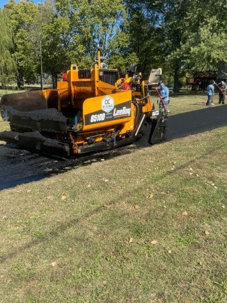 Yellow asphalt paving machine laying new pavement on a grassy area, with two workers in the background.