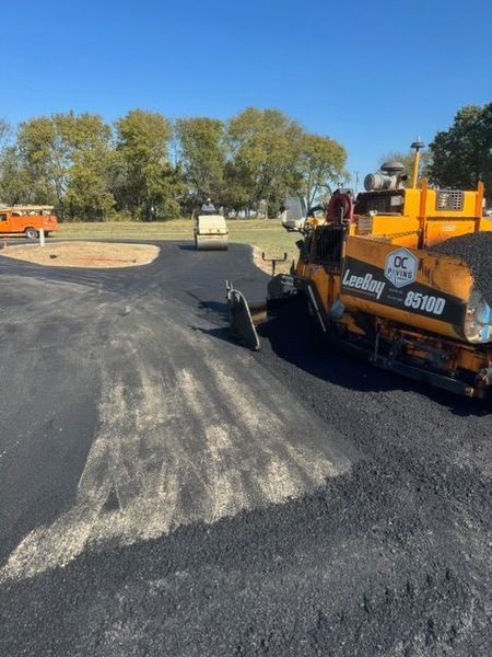 Asphalt paving in progress; black asphalt, yellow paver, blue sky, trees in the background.
