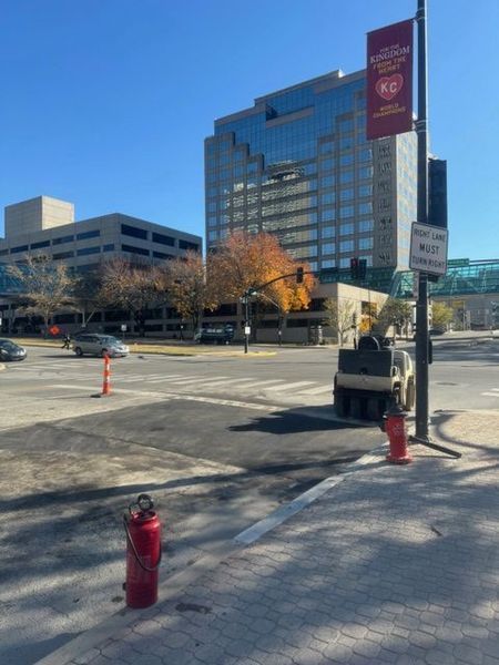 Asphalt roadwork in progress; steamroller on new black asphalt, red fire hydrant in foreground, city buildings in background.
