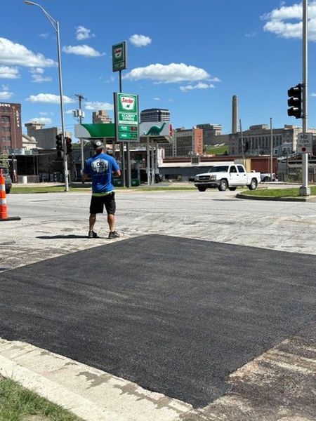 Man in blue shirt stands near newly paved asphalt patch in a city intersection with traffic and buildings.