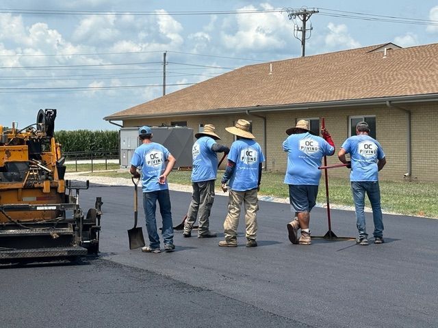 Paving crew laying asphalt on a road; workers wearing blue shirts and hats, building in the background.