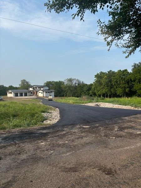 Asphalt road leading to a light-colored building under a partly cloudy sky, with green trees.
