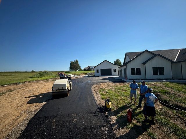 Asphalt driveway being laid in front of a modern white house and garage on a sunny day. Workers and a roller are present.
