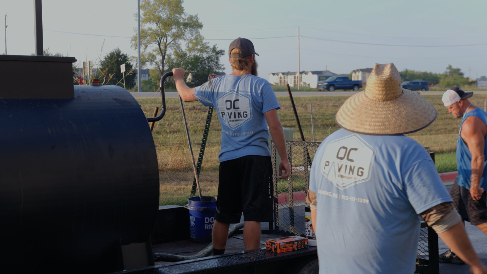 Three workers in light blue OC Paving shirts working with a large black asphalt trailer on a sunny day.
