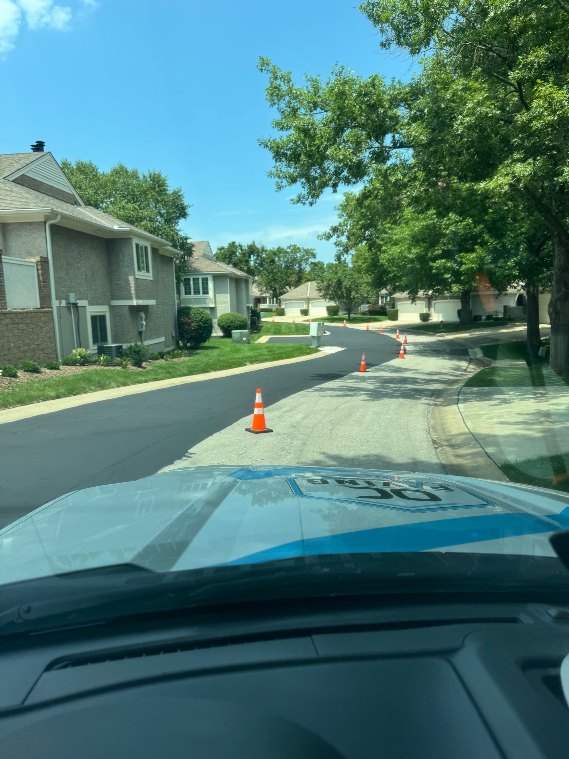 A view from inside a vehicle looking down a residential street with orange traffic cones marking fresh blacktop paving.