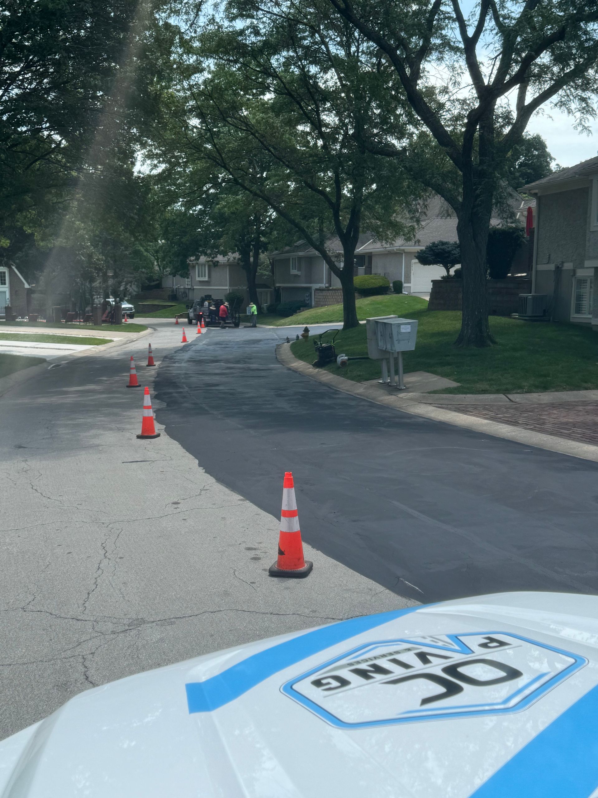 A freshly paved dark asphalt road marked by a row of orange traffic cones, viewed from a vehicle with a paving company logo.