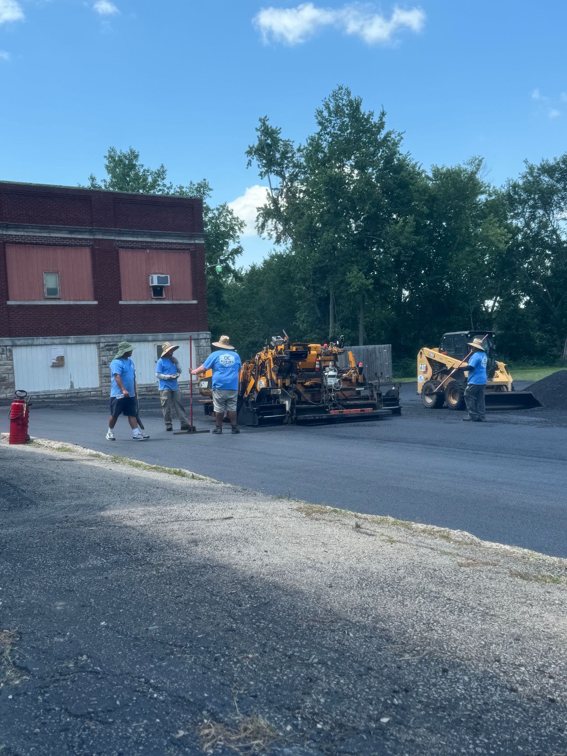 A crew in blue shirts operates paving machinery on a black asphalt surface next to a red brick building.