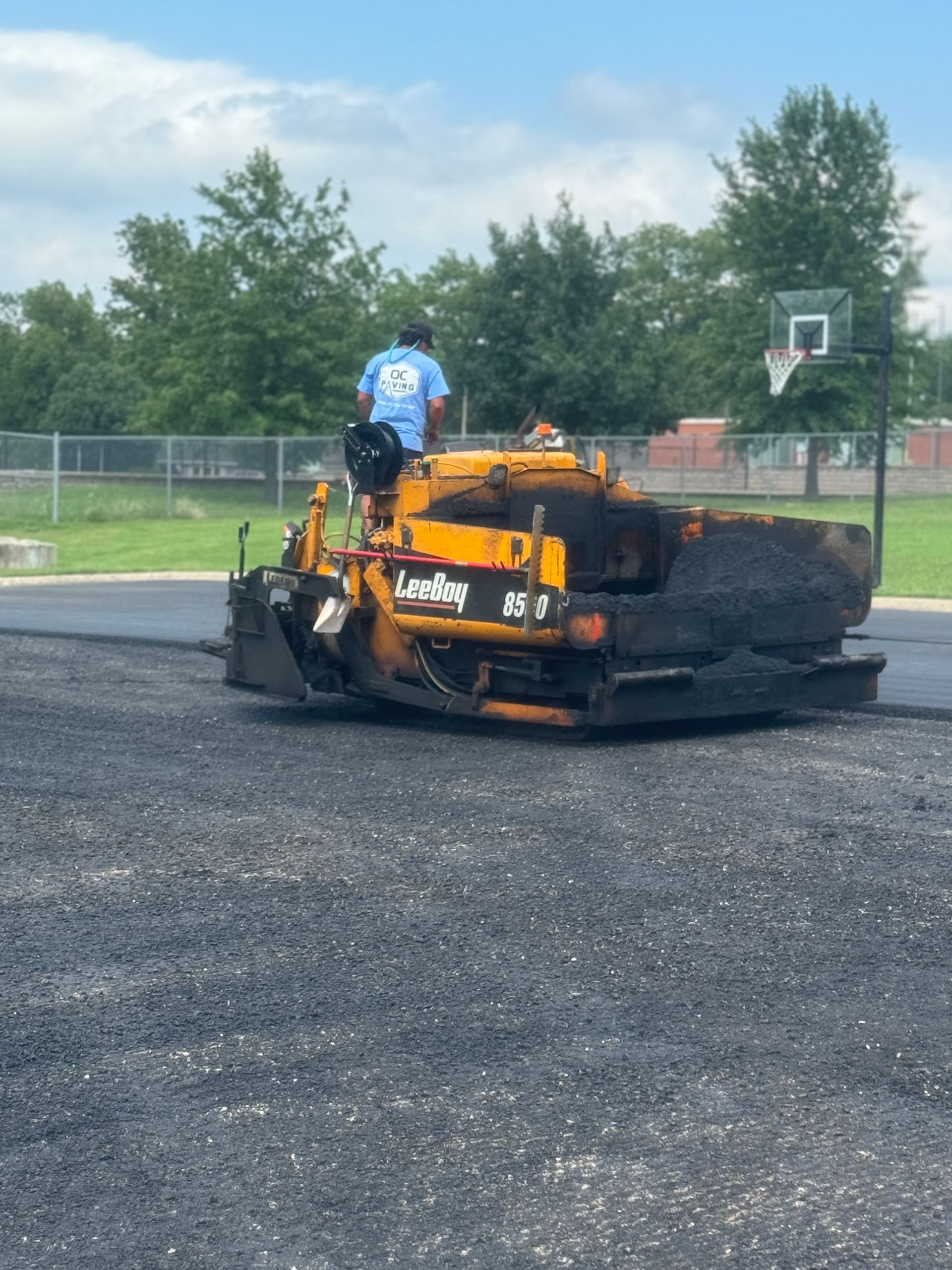 A construction worker operates a yellow asphalt paver on a paved surface near a basketball hoop and fence.