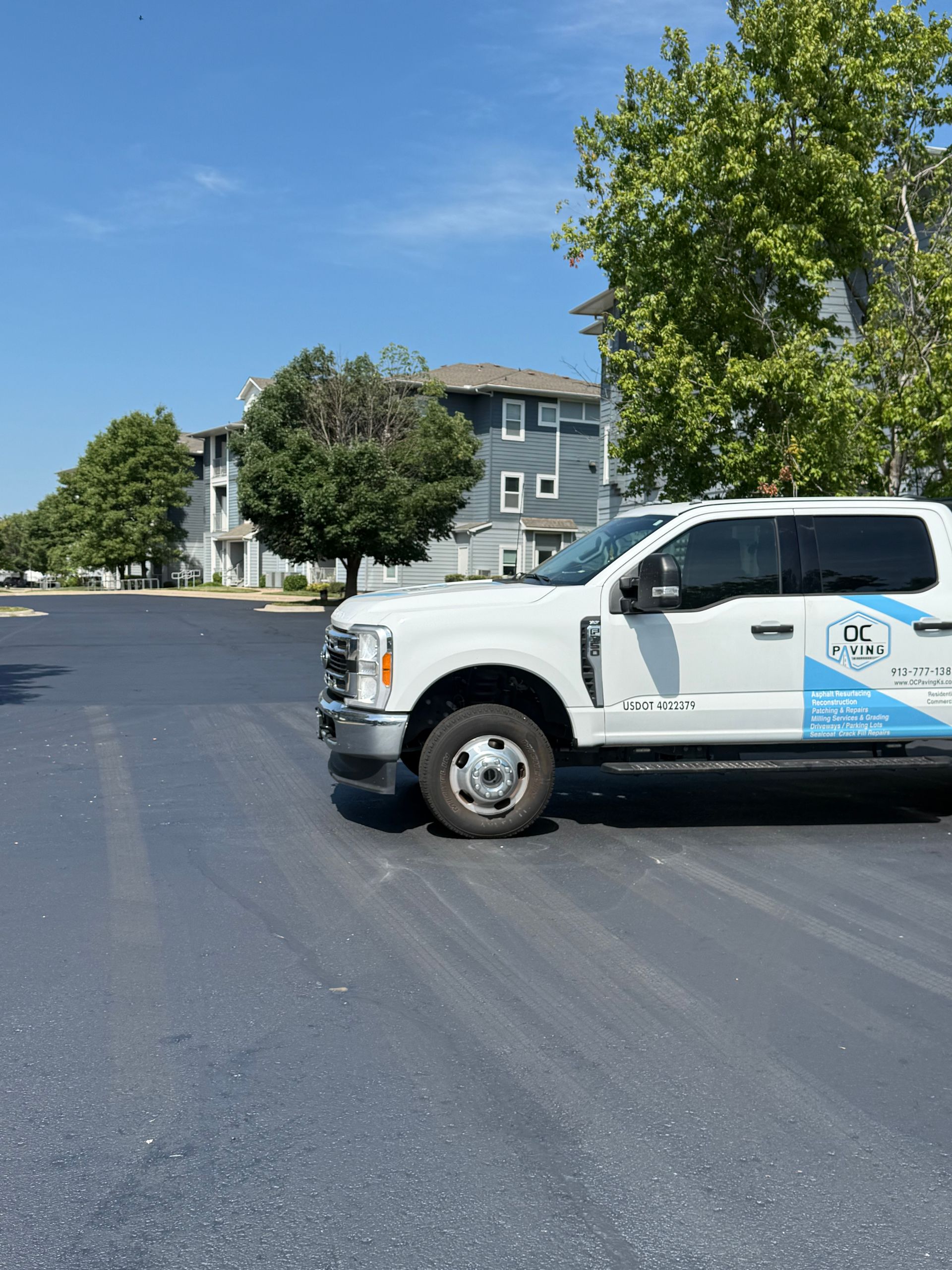 A white work truck parked on a freshly paved dark asphalt lot in front of a multi-story residential building.